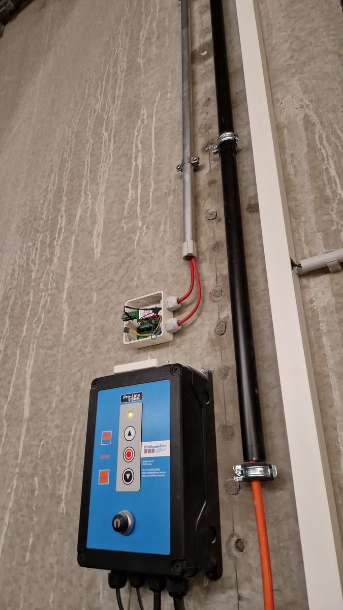 Torsteuerung an einer Betonwand, daneben eine geöffnete Abzweigdose mit dem Koppelmodul; eine rote Brandmeldeleitung führt im Rohr zur Steuerung