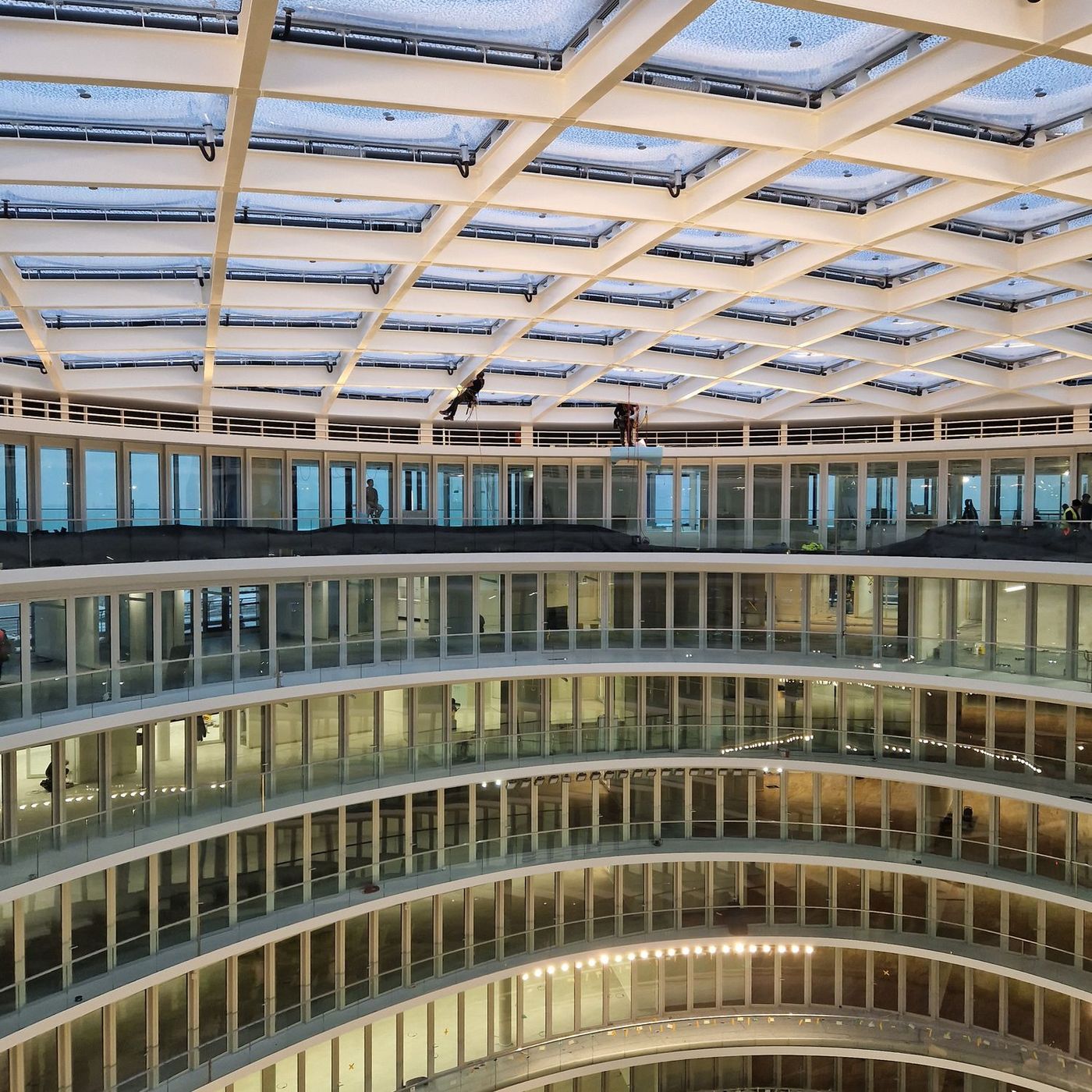 Zwei Höhenarbeiter am Seil unter dem gläsernen Rautendach eines mehrgeschossigen Atriums, darunter die umlaufenden Galerien