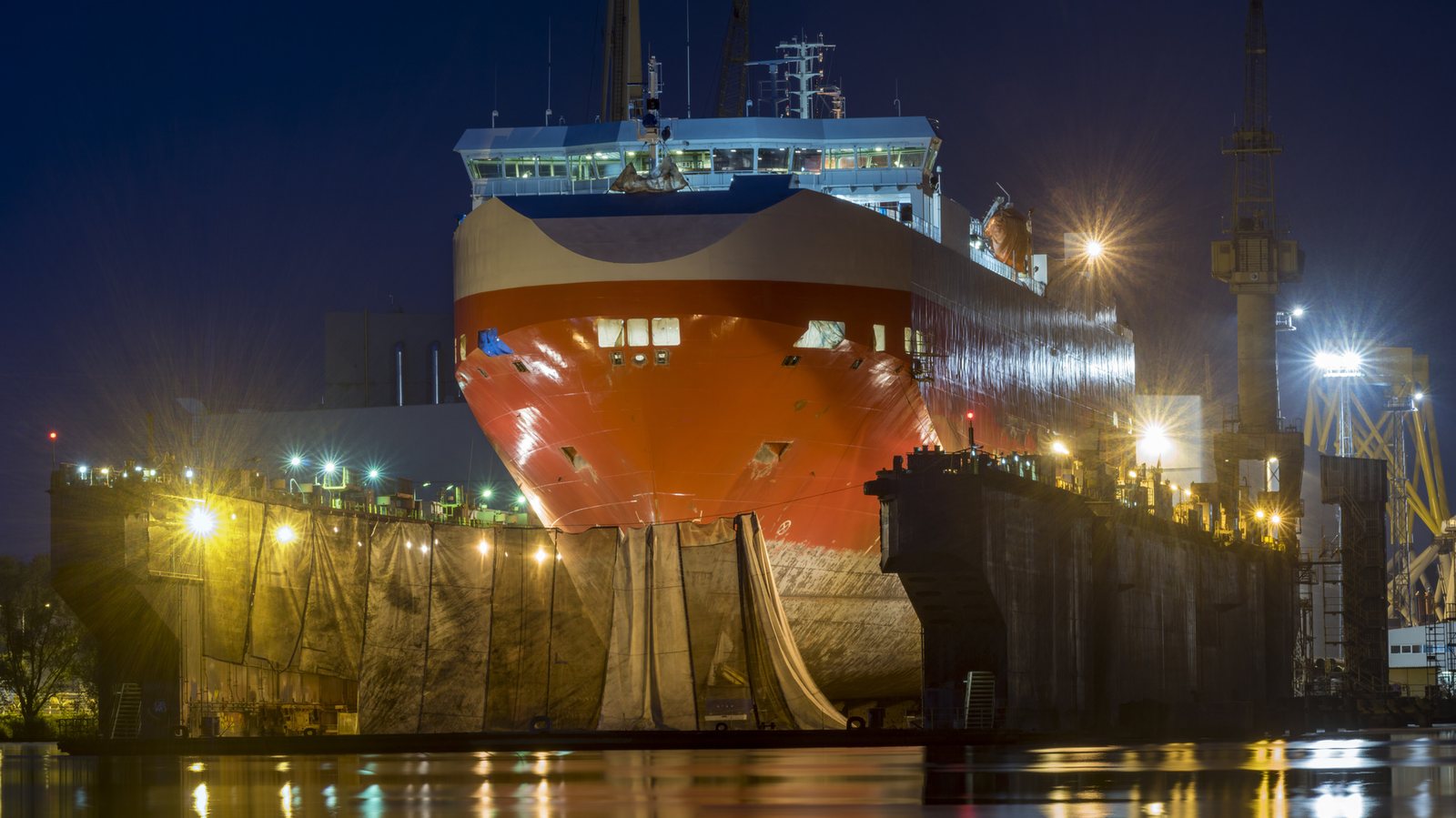 Schiffsneubau im Trockendock bei Nacht: der Rumpf steht auf Kielblöcken, die Werft ringsum ist ausgeleuchtet