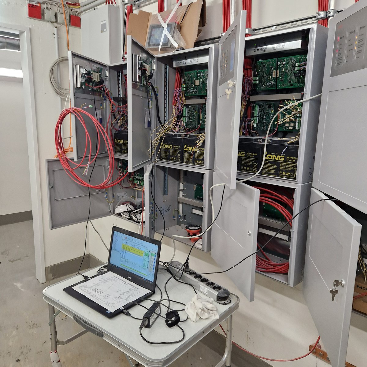 Reihe geöffneter Brandmelderzentralen mit Platinen, Notstromakkus und aufgelegten Ringleitungen; davor ein Laptop auf einem Klapptisch mit der Programmiersoftware